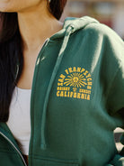 A person wearing the Hippie Hill Zip-Up Hoodie, a green cotton hoodie with yellow text saying "San Francisco Haight Ashbury California" and adorned with a sun graphic. Ideal for walks around Hippie Hill, this comfortable garment is crafted from 100% cotton.