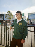 A person wearing the Hippie Hill Zip-Up Hoodie and a beanie smiles by a fence in a vibrant urban setting, capturing the relaxed atmosphere of Hippie Hill.