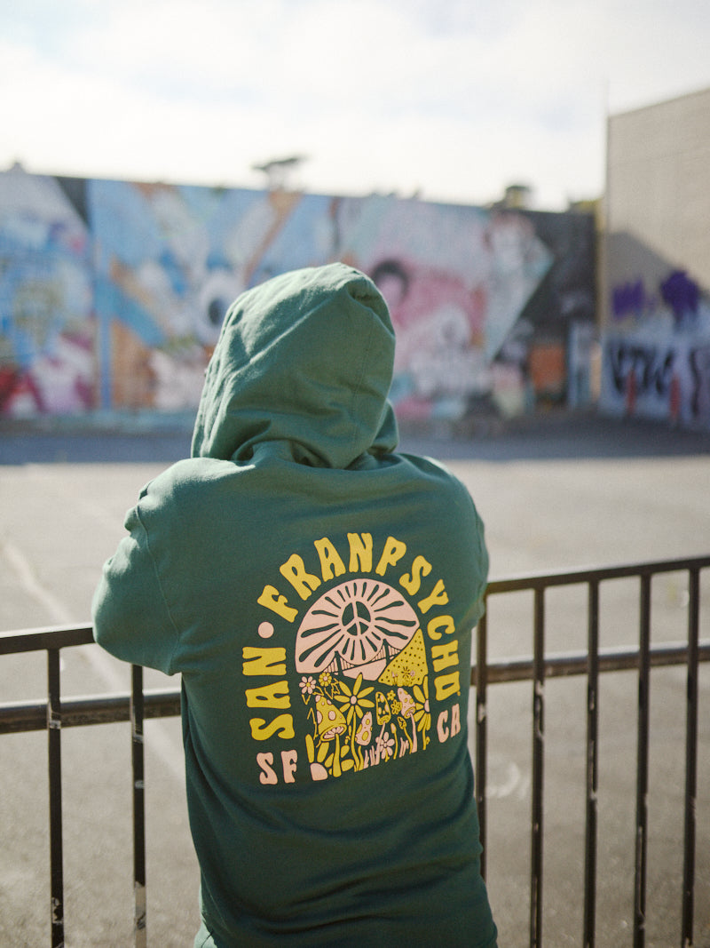 A person in the Hippie Hill Zip-Up Hoodie, made from 100% cotton and featuring a green design, stands against a backdrop of colorful street art, capturing the vibrant vibes of Hippie Hill.