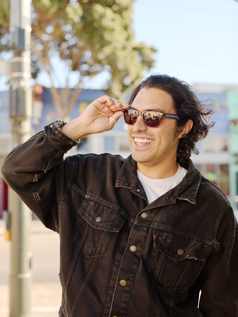 A smiling person in a denim jacket and SFP Sunglasses stands outdoors on a sunny day, enjoying the UV protection they provide.