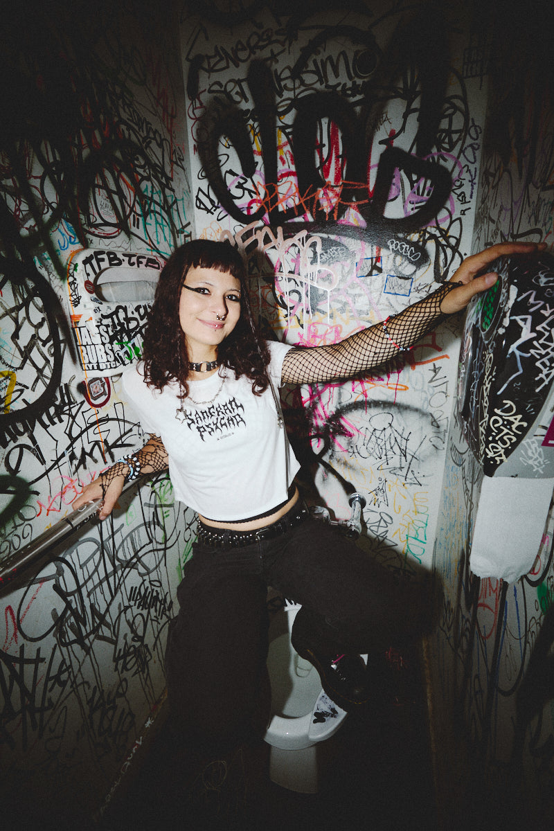 In a graffiti-adorned restroom, an individual with dark hair and piercings sits on a toilet, sporting a Metal Logo Crop Top from the Fall 2023 collection. The outfit is enhanced with fishnet sleeves, while the airlume cotton of the top provides a soft contrast to this daring style.