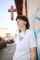 A person with piercings, wearing a Metal Logo Tee from the Fall 2023 collection, rests against a graffiti-covered wall under a Gospel Chapel sign on an urban street in San Francisco.