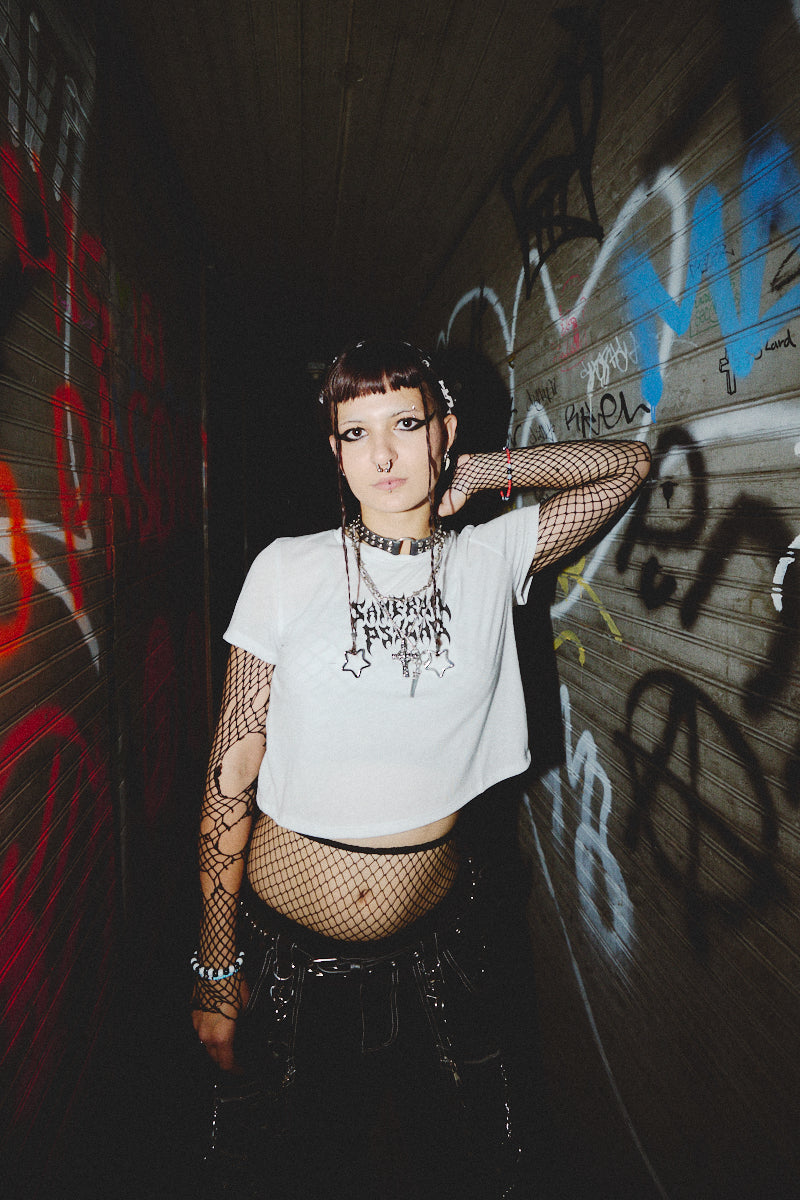 Dressed in the Metal Logo Crop Top from the Fall 2023 collection, an individual exudes confidence while showcasing its edgy design and mesh sleeves in a graffiti-filled hallway.