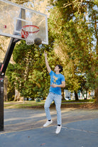 Dressed in a Live by the Three Blue Tee and jeans, made from soft cotton fabric, a person is playing basketball outdoors, leaping to shoot the ball like a true Warrior aiming for championships.