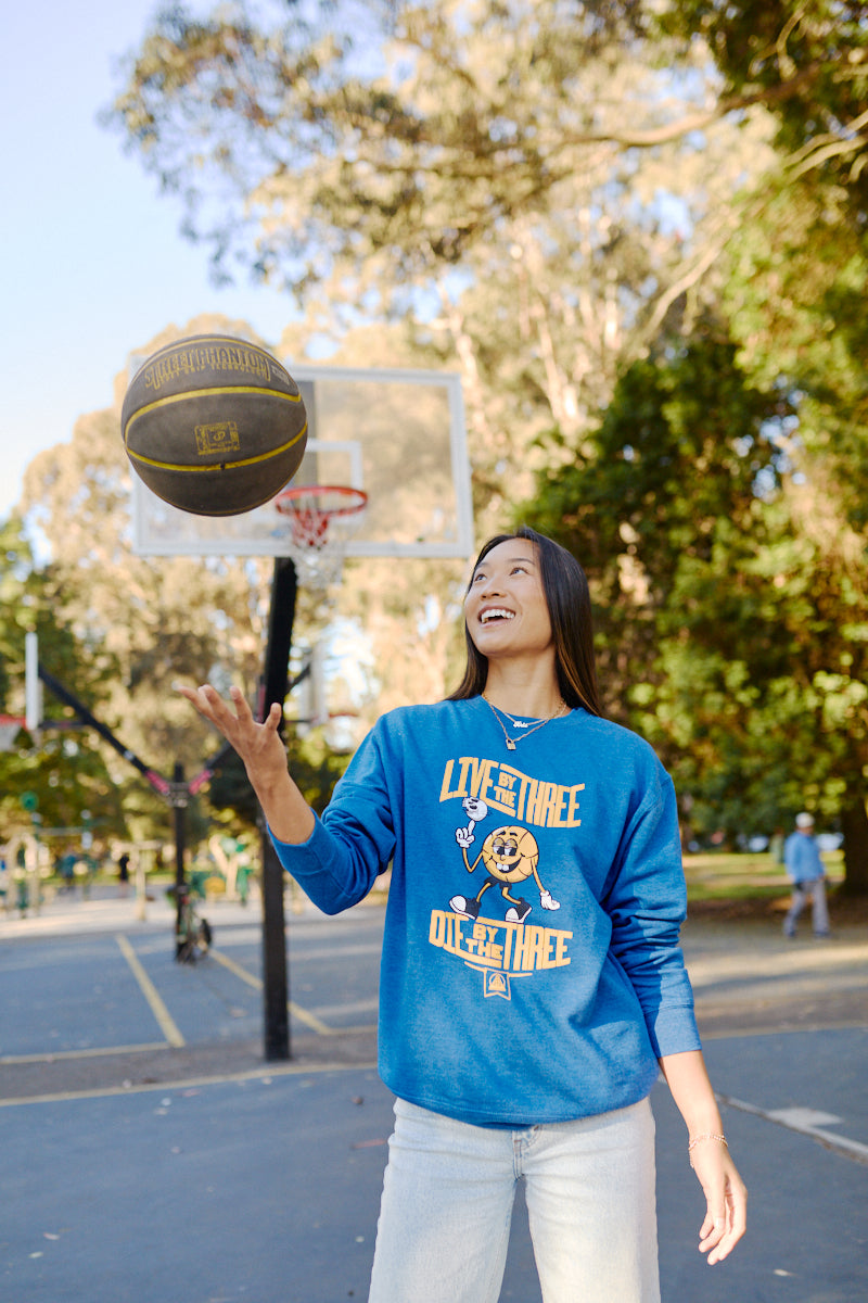 In a park, someone is skillfully spinning a basketball on their finger while wearing the Live by the Three Blue Crewneck.