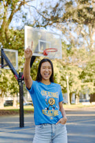 A smiling person wearing a "Live by the Three" Blue Tee raises one finger while standing on an outdoor basketball court, evoking the triumphant spirit of Warriors celebrating their latest championships.