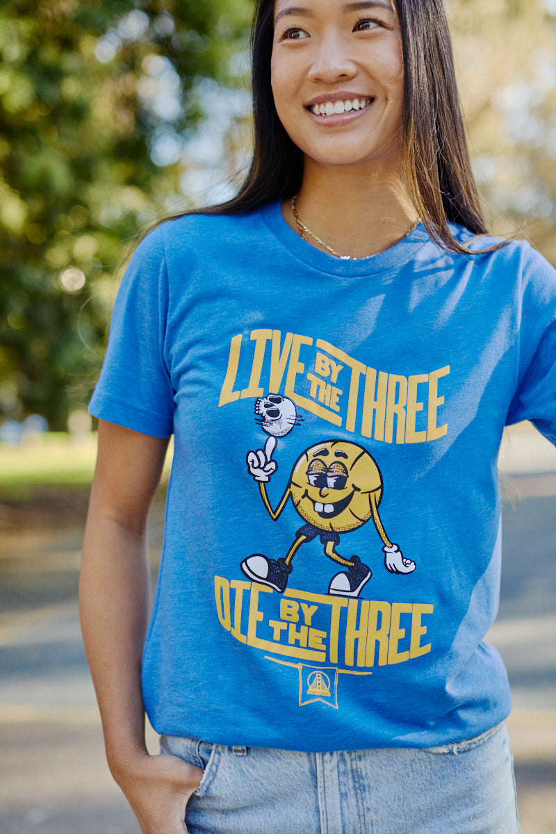 A person wearing the "Live by the Three Blue Tee," featuring a basketball graphic and this iconic slogan, stands out while proudly celebrating the Warriors' championships. The cotton fabrication guarantees both comfort and style on and off the court.