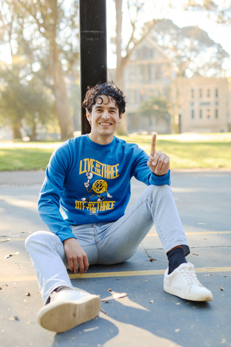 A smiling man sits on a basketball court, clad in a Live by the Three Blue Crewneck and jeans, pointing upward as if celebrating like the Warriors after winning championships.