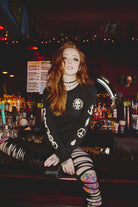 A woman with long red hair sits on a bar counter, wearing ripped leggings and the BBNW Skull Long Sleeve—a black tee that adds an edgy vibe to her look.