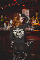 A woman with red hair sits at a bar, wearing the BBNW Skull Long Sleeve—a black shirt featuring hand-printed San Francisco punk-inspired skull graphics and Sail Tranpsycho text.