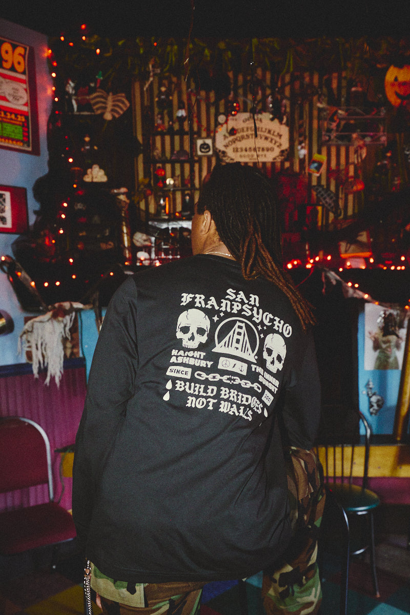 Wearing the BBNW Skull Long Sleeve, a person sits in a decorated, dimly lit, and colorful room, channeling punk shirt vibes with hand-printed San Francisco flair.