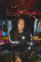A man with long dreadlocks sits on a chair in a colorful room adorned with string lights and eclectic wall decor, wearing the BBNW Skull Long Sleeve shirt.