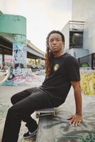 In a graffiti-covered skate park, a long-haired person sits on a skateboard, rocking the Banned in SF Tee. This scene embodies Fall Punk Rock 2023 in San Francisco, where street art meets rebellious fashion.