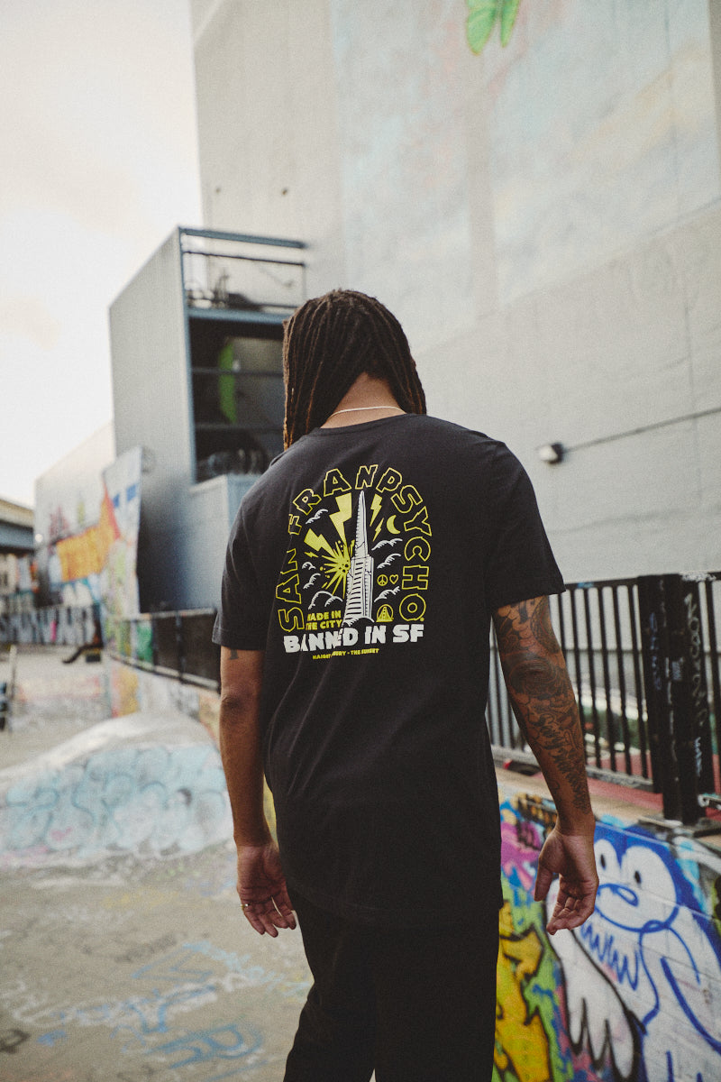 A person with dreadlocks, wearing the Banned in SF Tee, walks through a city neighborhood where graffiti-covered walls reflect the rebellious spirit of Fall Punk Rock 2023.