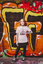 In front of a vibrant graffiti wall, a person with tattoos strikes a pose wearing a Shredder Logo Tee and black skirt.