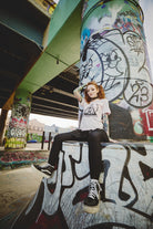 A tattooed individual sits on a graffiti-covered structure beneath a bridge, donning the Shredder Logo Tee from the Punk Rock 2023 collection along with sneakers.