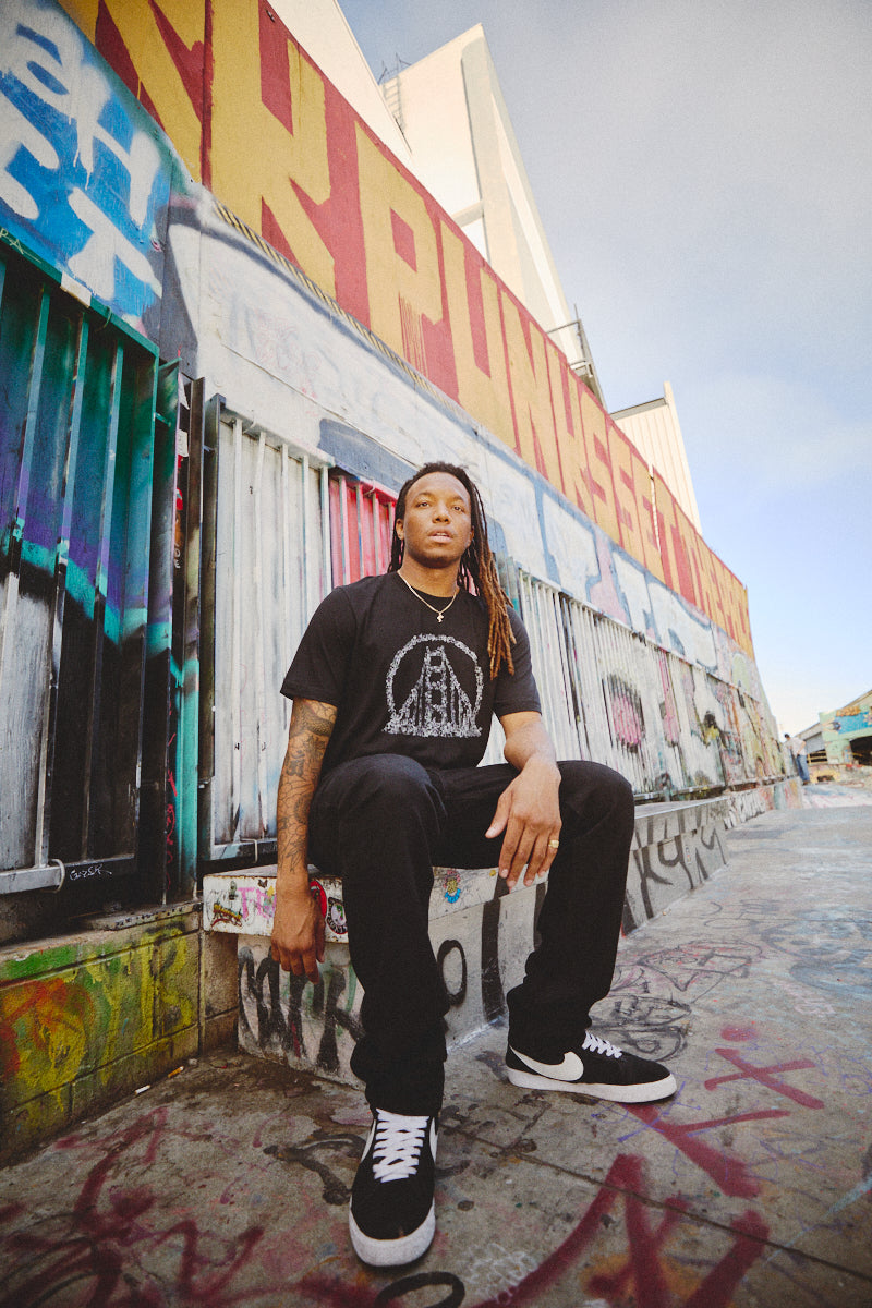 A person sits in front of a graffiti-covered wall, effortlessly showcasing a punk rock vibe with their fashionable Shatter Logo Tee and black sneakers.