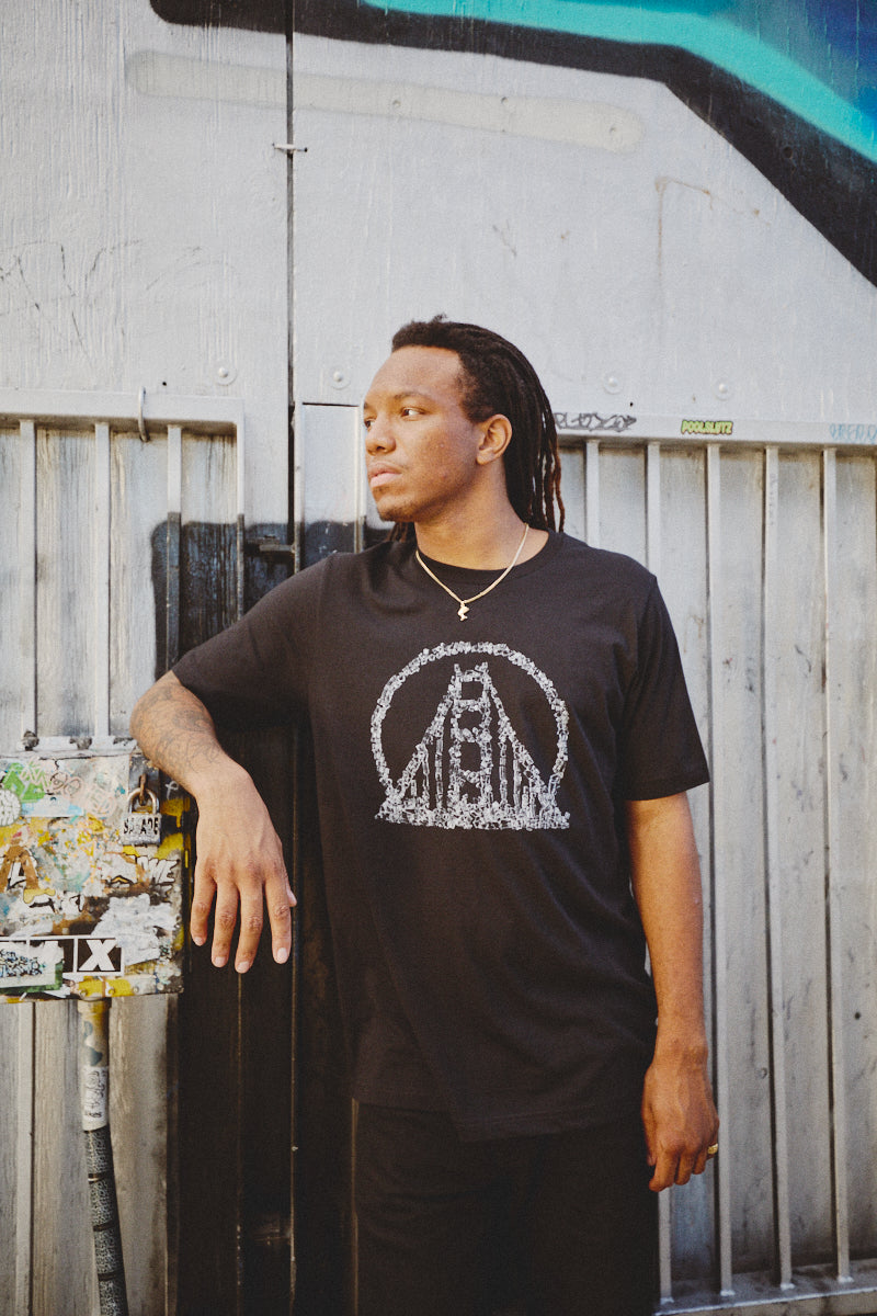 A person with dreadlocks wearing a Shatter Logo Tee leans against a wall in an urban setting, embodying the edgy vibe of Fall Punk Rock 2023.