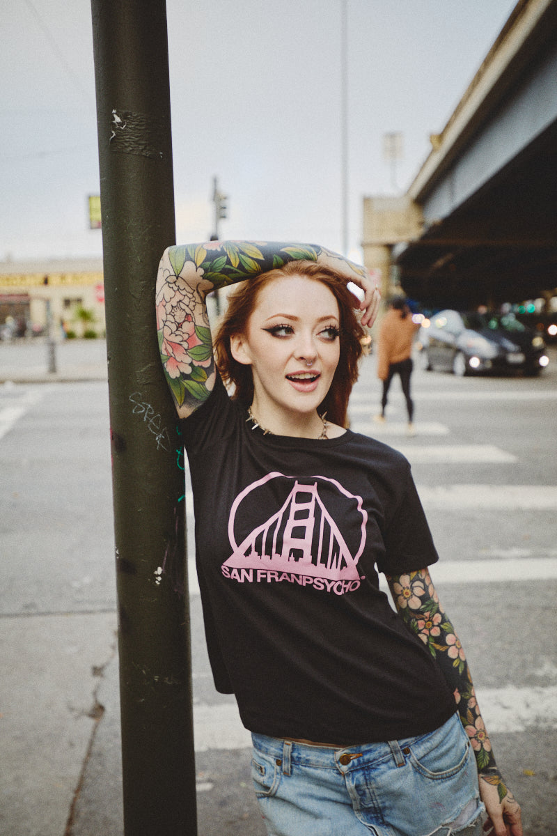 A woman wearing a Women’s Black & Pink Logo Tee featuring a vintage San Franpsycho Golden Gate Bridge graphic.