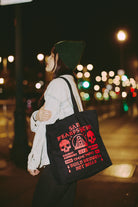A woman in a beanie and denim jacket holds the BBNW Skull Tote, featuring hand-printed red graphics and text, at night.