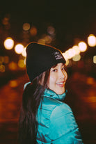 A person wearing a teal jacket and the Black Waffle Beanie w/ Wet & Wreckless smiles at night against blurred city lights, proudly showing off the patch.