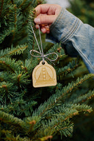 A hand hangs a SFP Wood Ornament, a wooden decoration shaped like the Golden Gate Bridge, on a Christmas tree. Laser-etched from bamboo, this exquisite ornament adds a unique touch of elegance and charm to the festive setting.