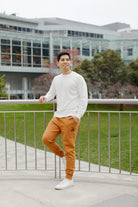 A man in a white shirt and brown pants leans on a railing outside a modern building, with the Adobe & Black Embroidered Logo Joggers featuring rib-knit ankle cuffs adding a touch of casual elegance.