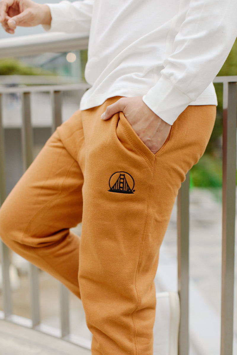 A person wearing Adobe & Black Embroidered Logo Joggers, with a bridge logo and hand in pocket, stands near a metal railing outdoors. The joggers feature rib-knit ankle cuffs for style and comfort.