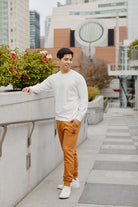 A smiling man in a white shirt and Adobe & Black Embroidered Logo Joggers, featuring rib-knit ankle cuffs, stands on an urban walkway with plants and buildings in the background.