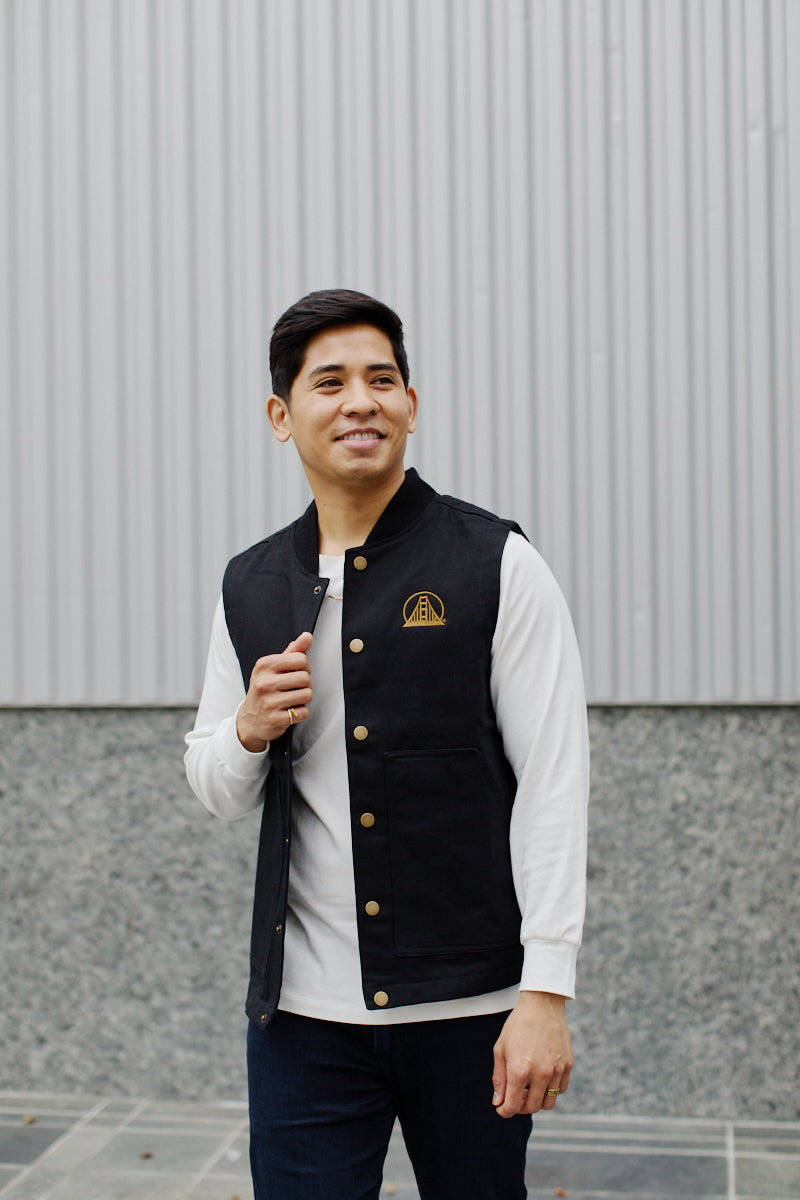 A person wearing an Embroidered Logo Workwear Vest in black, paired with a white shirt, stands smiling in front of a gray corrugated wall while looking to the side.