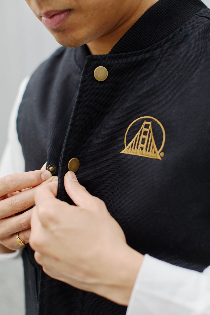 A person is wearing the Embroidered Logo Workwear Vest, a black jacket adorned with gold buttons and an embroidered bridge logo, fastening the buttons. Made from 100% cotton canvas fabric, this piece combines durability with classic workwear style.