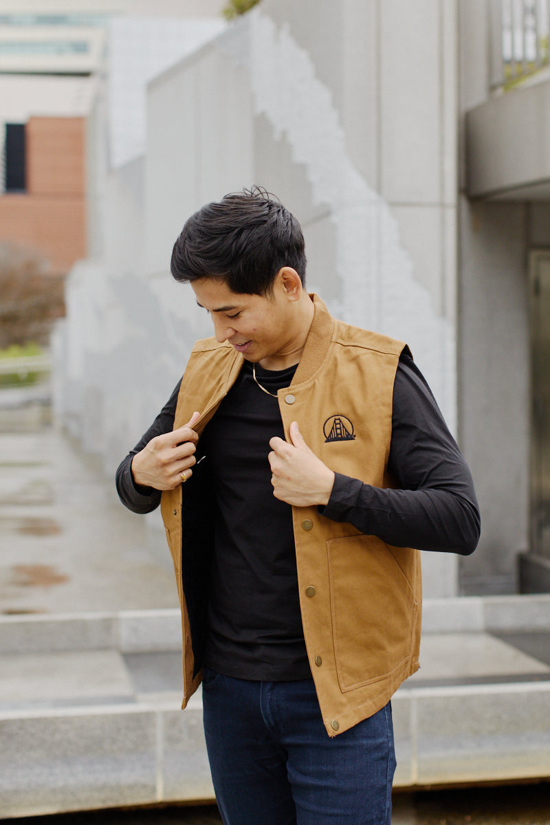 Standing outdoors on a bustling city street, a man wearing a black shirt adjusts his Embroidered Logo Workwear Vest, made from 100% cotton canvas.