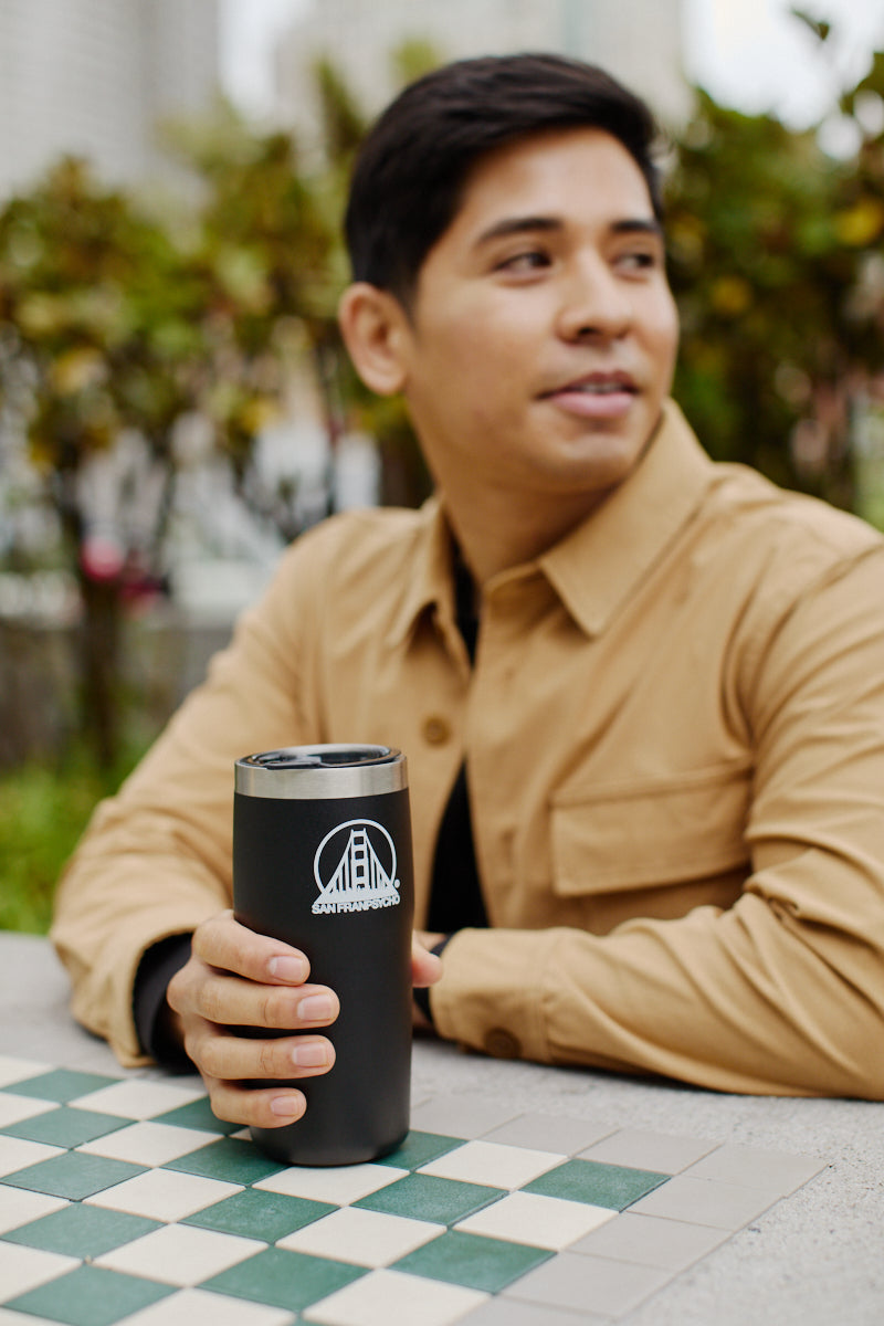 A man in a tan jacket holds an SFP Logo Tumbler, sitting outdoors at a checkered table, gazing thoughtfully to the side.