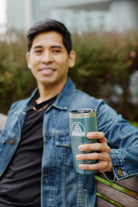 A person in a denim jacket holds an SFP Logo Tumbler while sitting on a bench outside, savoring the moment.