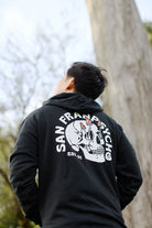 The person stands outdoors wearing a Shroom Skull Pullover Hoodie made from 100% cotton, with the design displayed on the back, looking up.