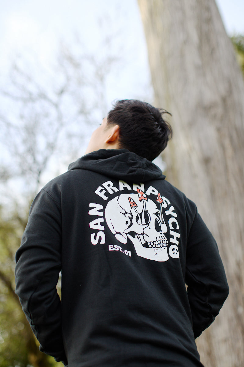 The person stands outdoors wearing a Shroom Skull Pullover Hoodie made from 100% cotton, with the design displayed on the back, looking up.