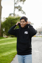 A smiling man in a 100% cotton black hoodie stands on a park path with trees in the background, looking as relaxed as if he were wearing the Shroom Skull Pullover Hoodie.