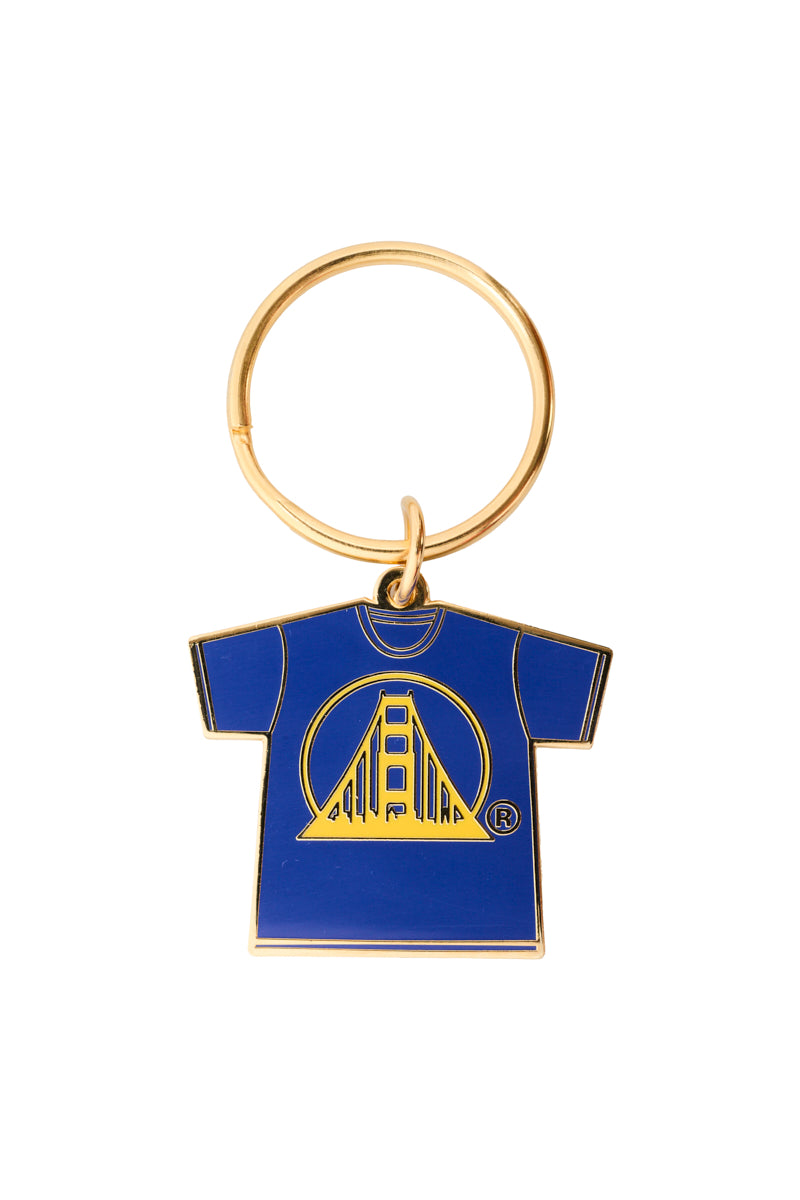The Logo Tee Keychain showcases a metal keychain with a blue t-shirt charm adorned by a yellow bridge design and vibrant enamel filling.