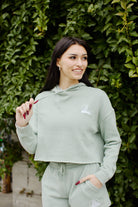 A smiling woman in a green tracksuit stands in front of a leafy green background, her sage Embroidered Logo Crop Hoodie with the San Franpsycho logo showcasing her relaxed fit.