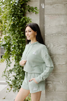 A woman in an Embroidered Logo Crop Hoodie with a relaxed fit stands by a leafy wall, smiling outdoors.