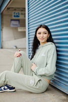 A young woman wearing Women's Embroidered Logo Joggers and a green tracksuit with rib-knit cuffs sits by a blue garage door, holding a lollipop and gazing to the side.