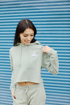 A woman in an Embroidered Logo Crop Hoodie and relaxed fit sweatpants poses in front of a blue shuttered background, smiling and looking down.