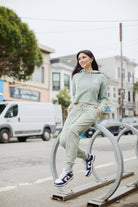 A person in a green hoodie and Women's Embroidered Logo Joggers sits on a bike rack, with buildings and a white van in the background.