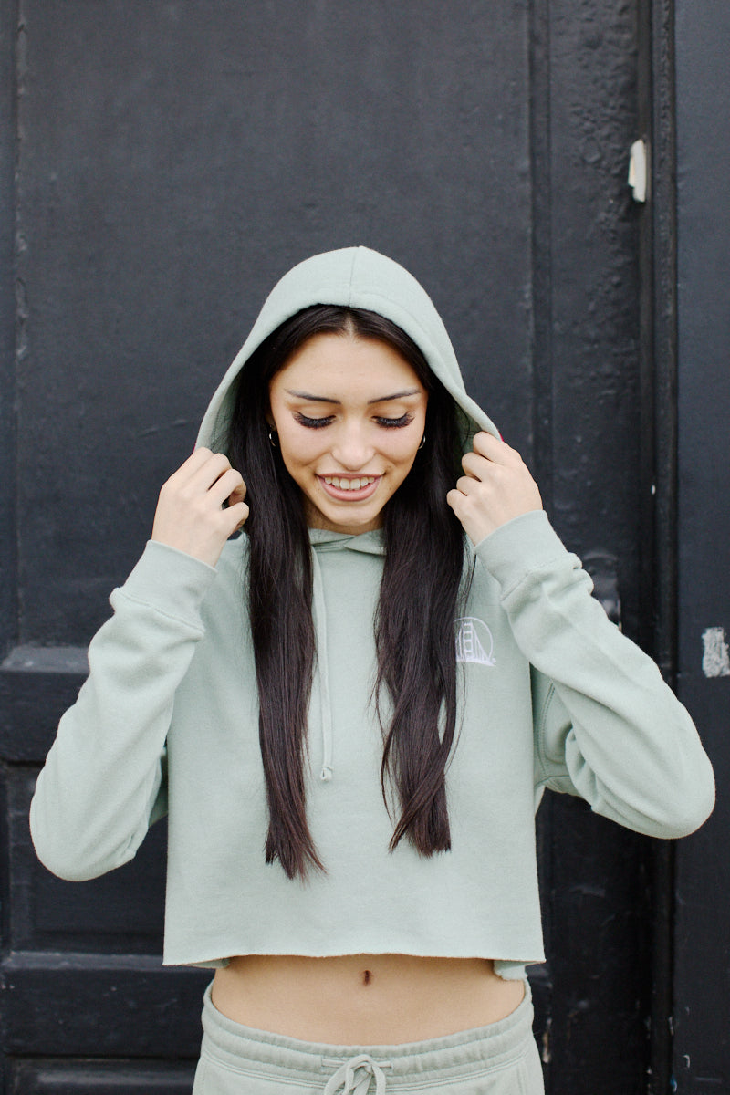 A person wearing the Embroidered Logo Crop Hoodie in sage, adorned with the San Franpsycho logo, smiles while adjusting the hood against a dark background.