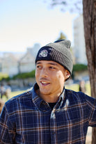 Wearing the Black Waffle Beanie w/ Shroom Skull, a man in a plaid shirt stands outdoors by a tree, looking to the side and smiling—a laid-back nod to San Franpsycho style.