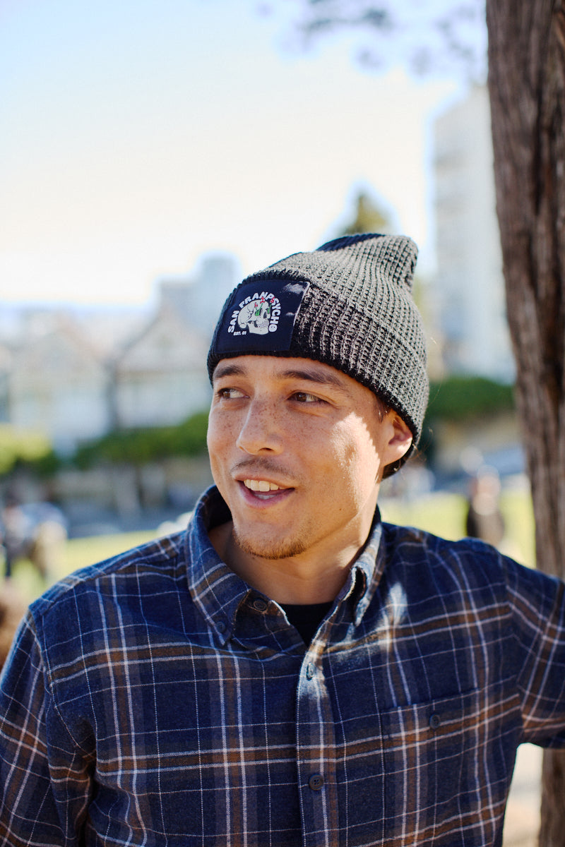 Wearing the Black Waffle Beanie w/ Shroom Skull, a man in a plaid shirt stands outdoors by a tree, looking to the side and smiling—a laid-back nod to San Franpsycho style.
