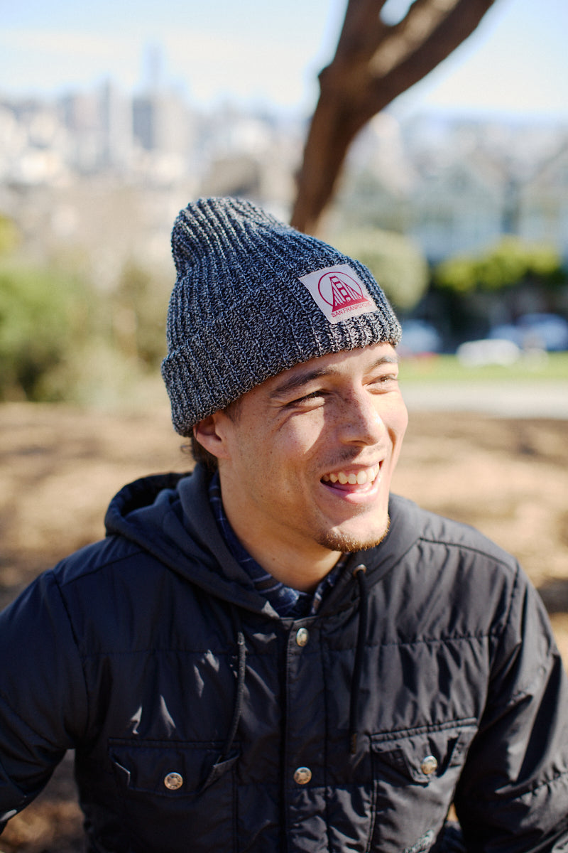 Smiling person outdoors on a sunny day wearing a Zebra Lumberjack Beanie with a Natural/Red SFP Logo and a black jacket.