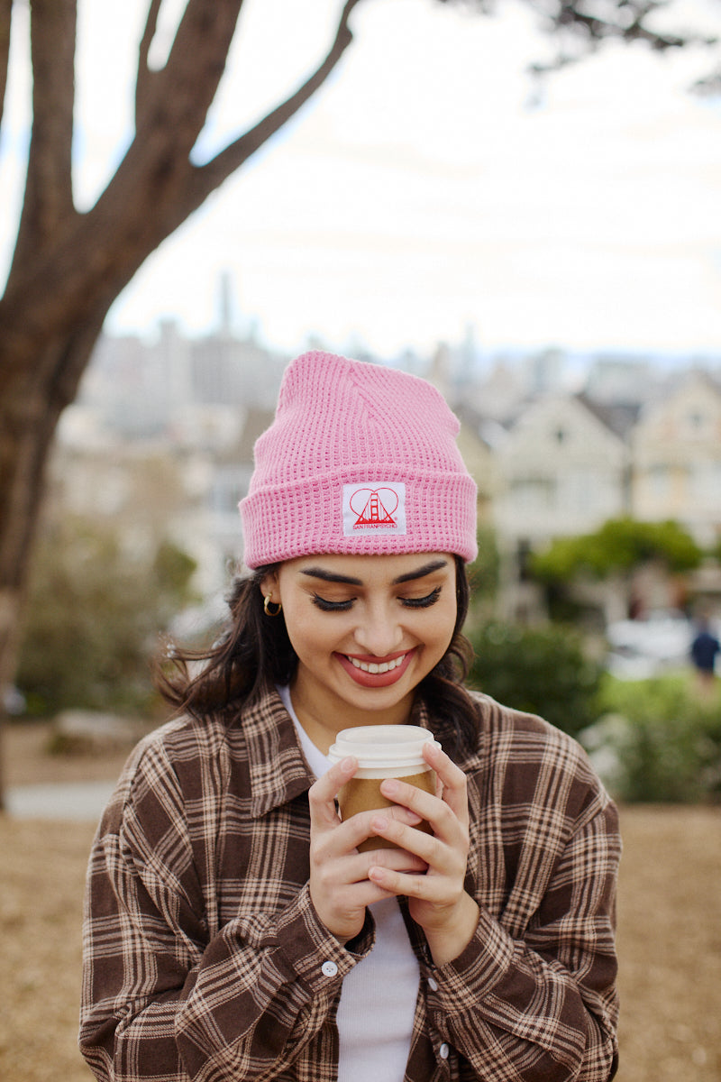 A smiling person outdoors holds a coffee cup against the backdrop of trees and the San Franpsycho cityscape, wearing a Pink Waffle Beanie w/ Heart Logo that adds charm to the vibrant scene.