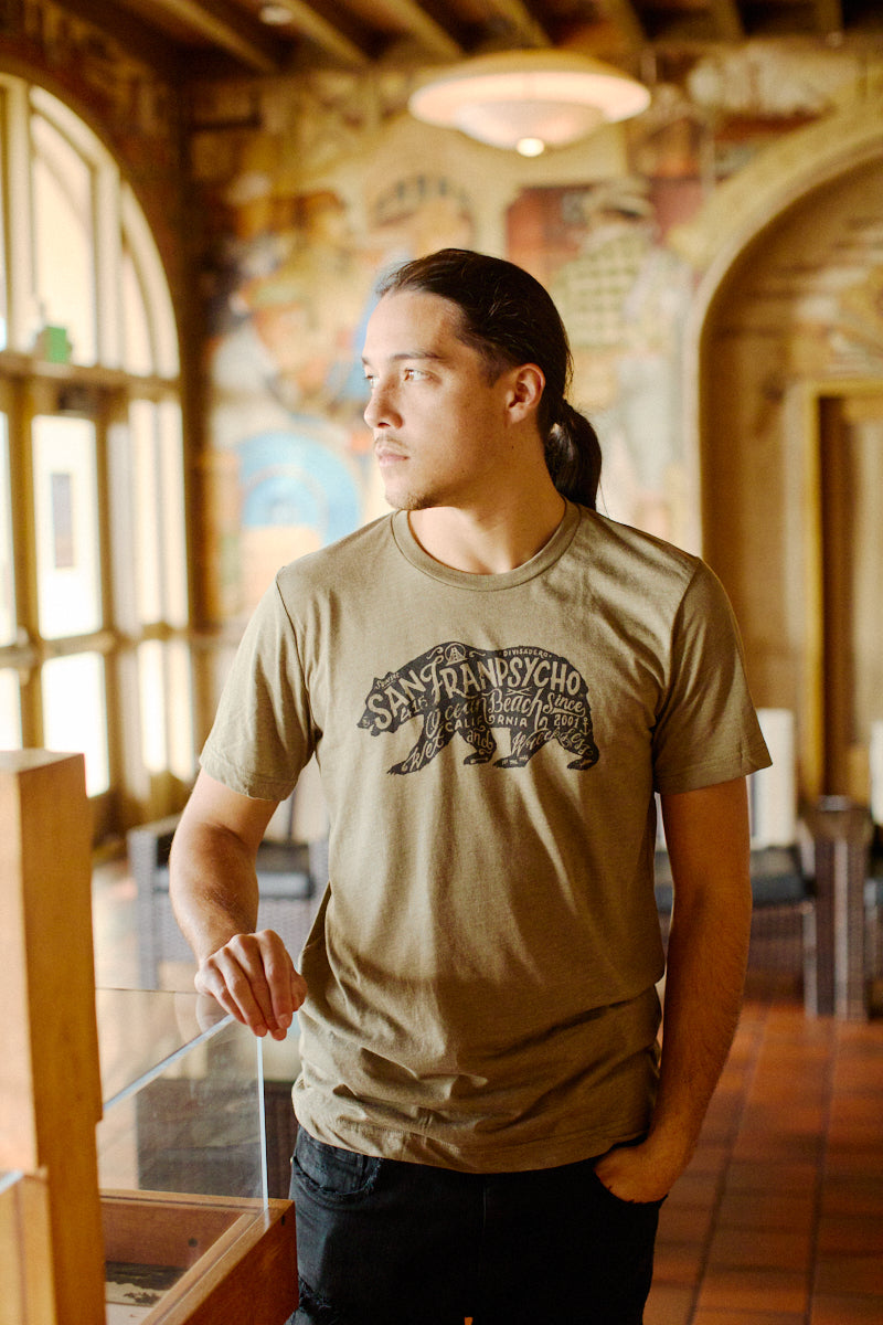 A man with long hair in a ponytail wears the Cali Bear Tee from San Franpsycho, standing indoors and looking to the side—a signature Kevin Espeche style.
