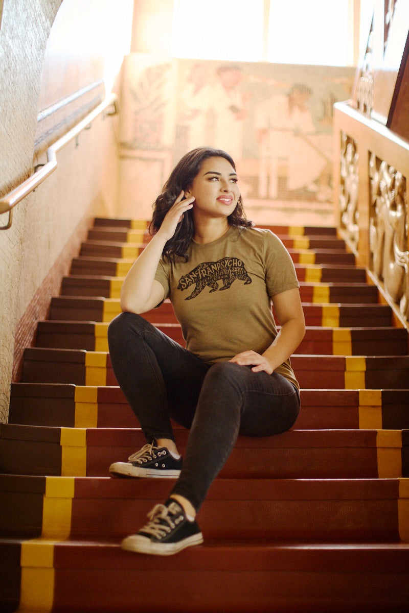 Smiling woman in the Cali Bear Tee sits on colorful stairs in a sunlit room, embodying a relaxed vibe and looking to the side.